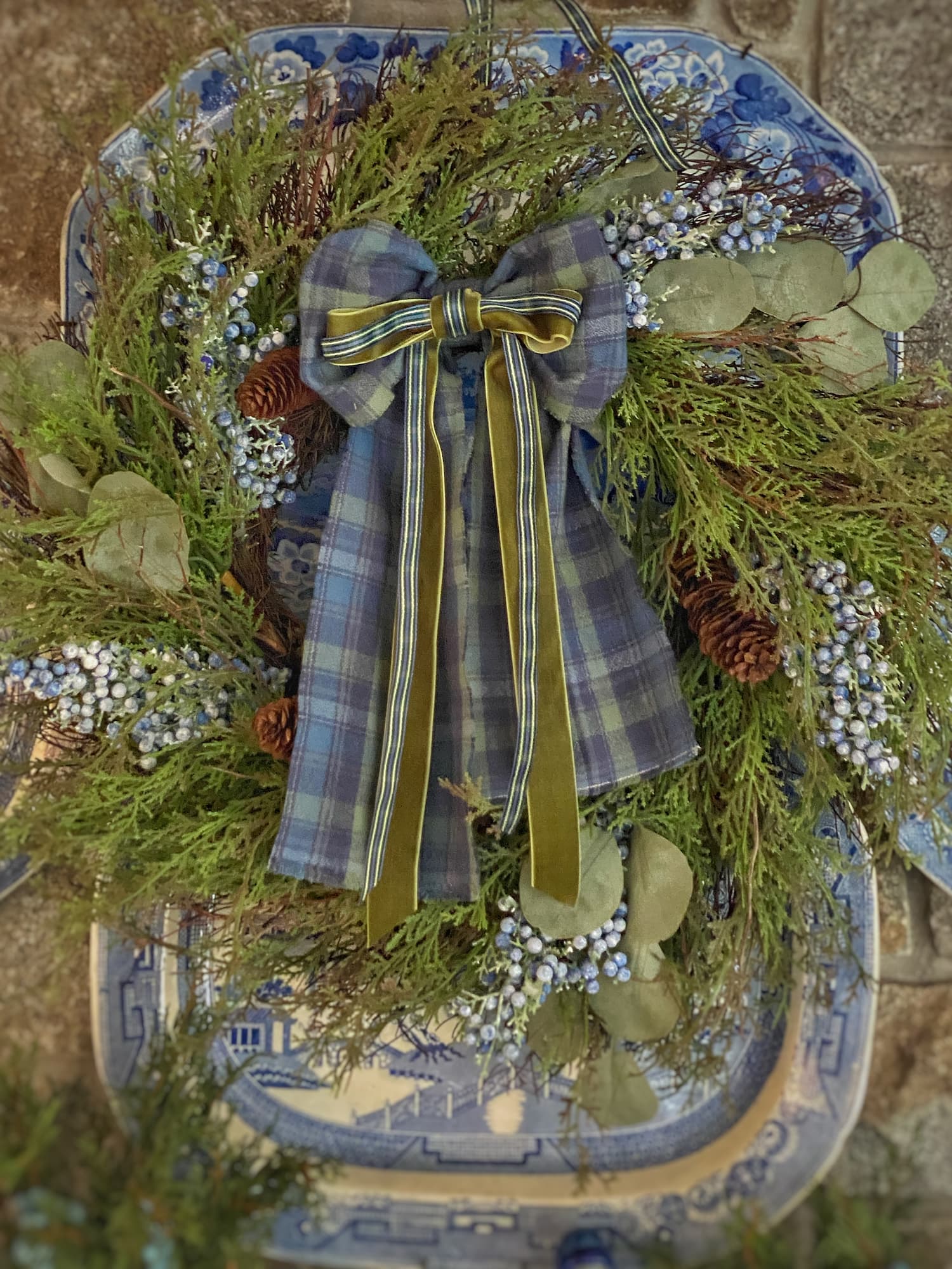 A holiday wreath made of faux juniper with blue berries, pinecones, and a large plaid flannel bow layered with green velvet ribbon, displayed on a vintage blue-and-white transferware platter.