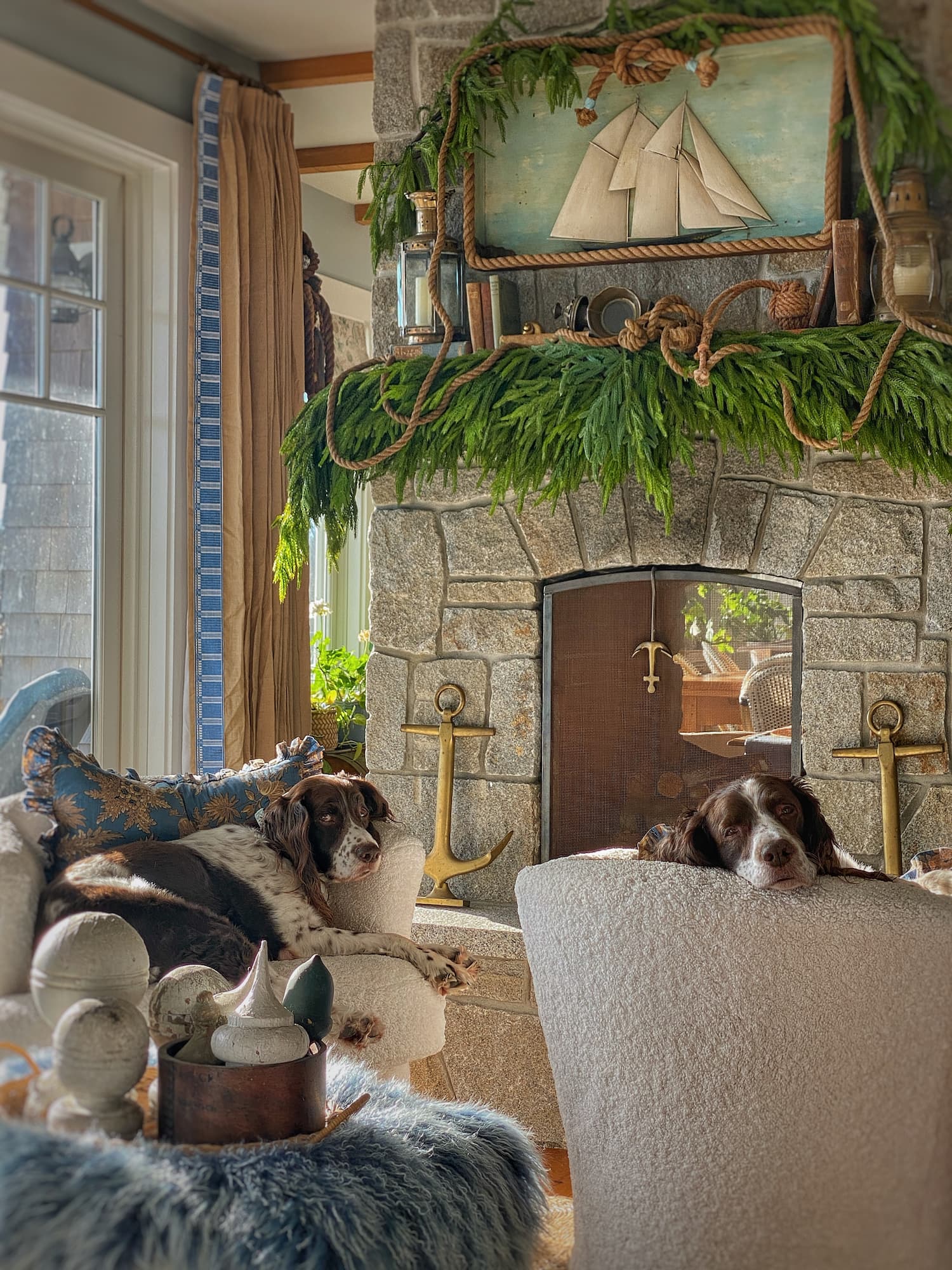 Two Springer Spaniels lounging in cozy chairs in front of a stone fireplace decorated with coastal greenery, nautical rope, and a vintage ship diorama in a Maine seaside cottage.
