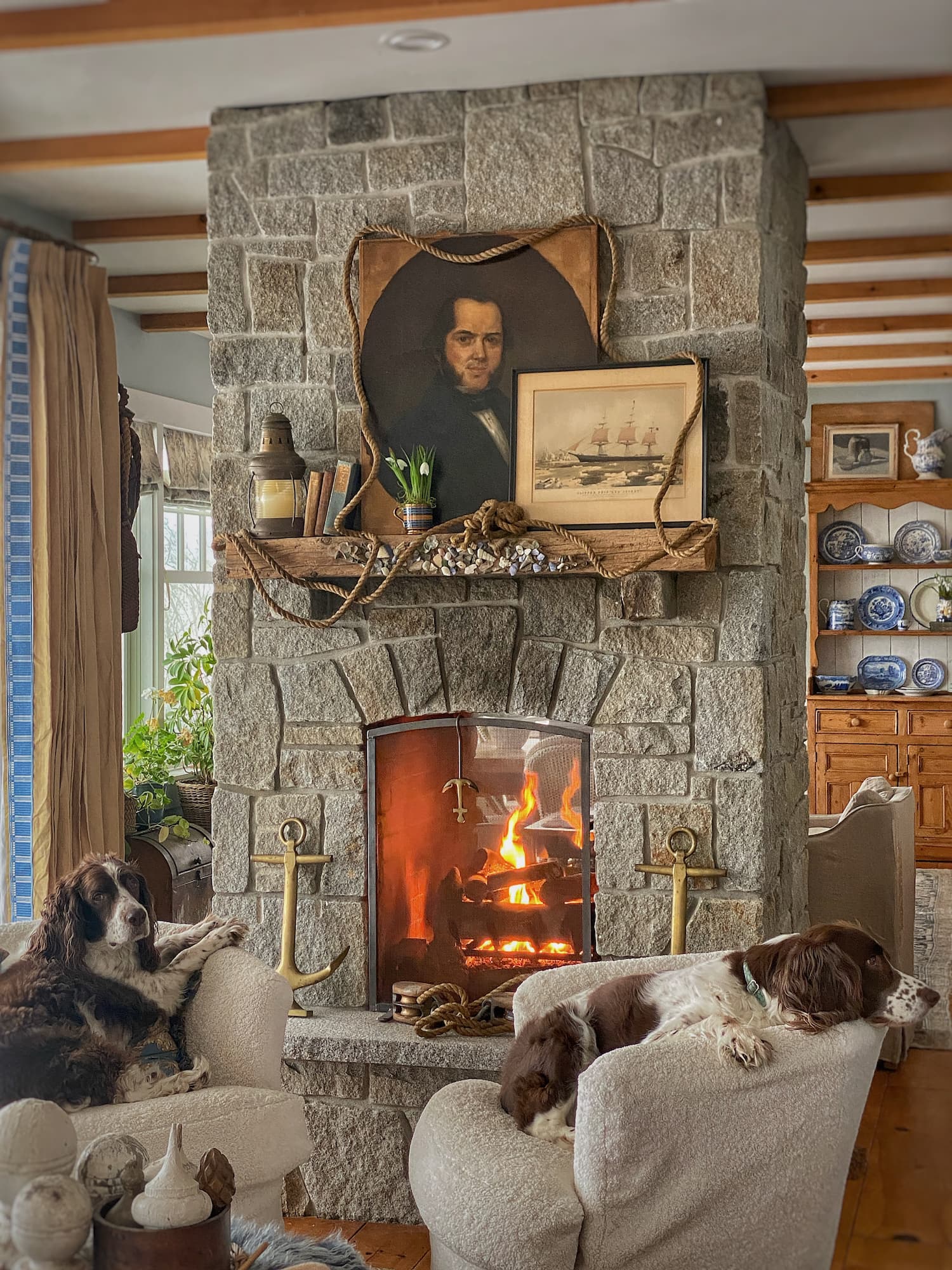 Stone fireplace with a nautical mantel featuring a vintage portrait, ship print, rope accents, and faux snowdrops, with two brown-and-white Springer Spaniels resting in cozy white chairs by the fire.
