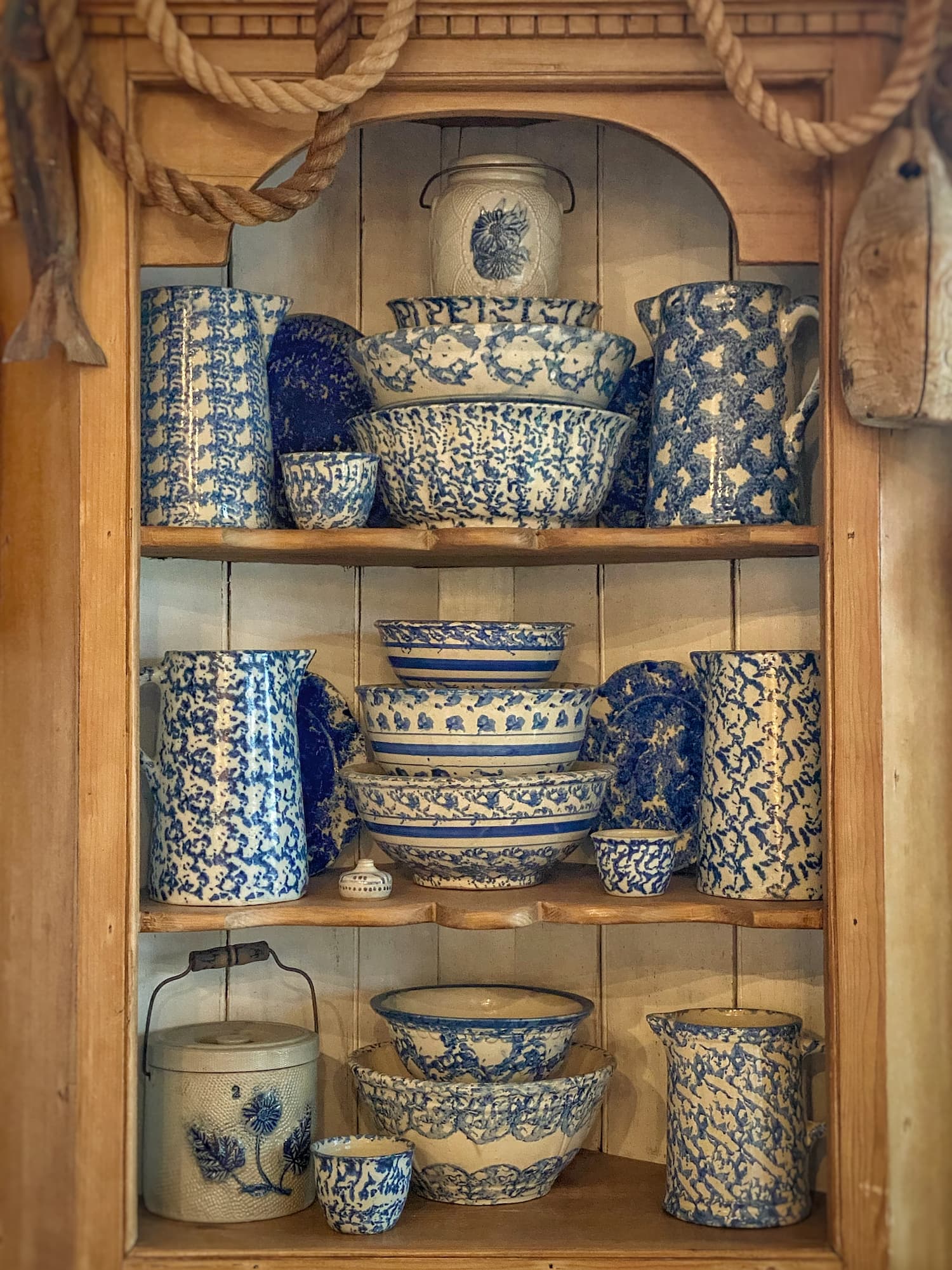 Blue and white spongeware bowls and pitchers displayed on shelves in a wooden corner hutch in a Maine cottage dining area.