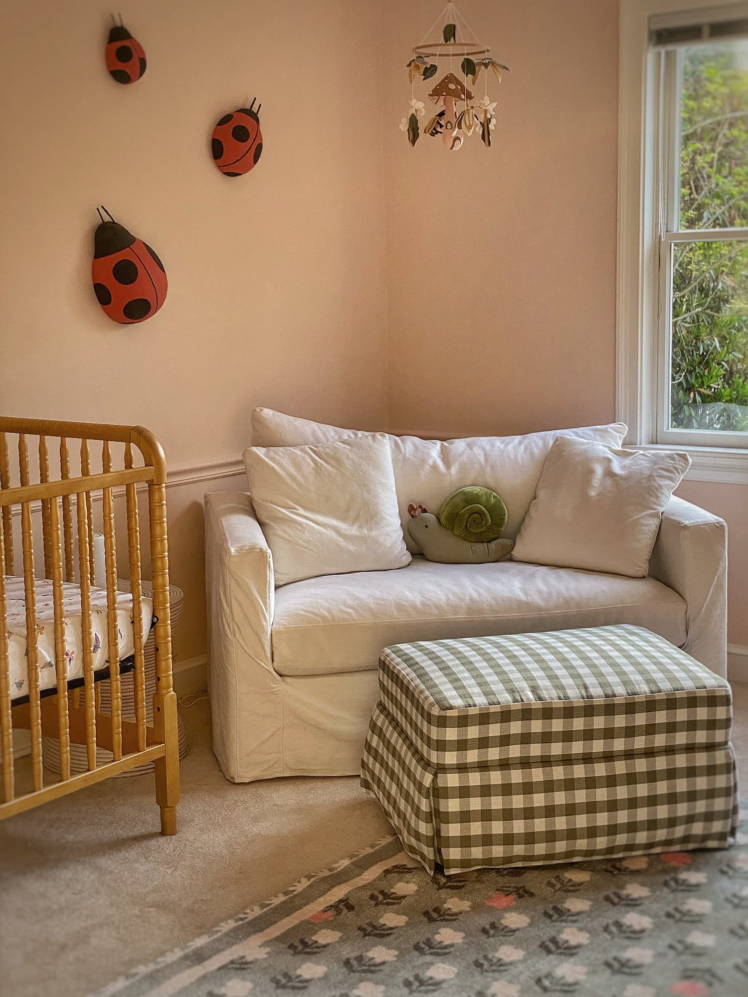Pink nursery with Benjamin Moore Tissue Pink walls, ladybug wall decor, wood crib, slipcovered loveseat, floral rug, and green gingham ottoman