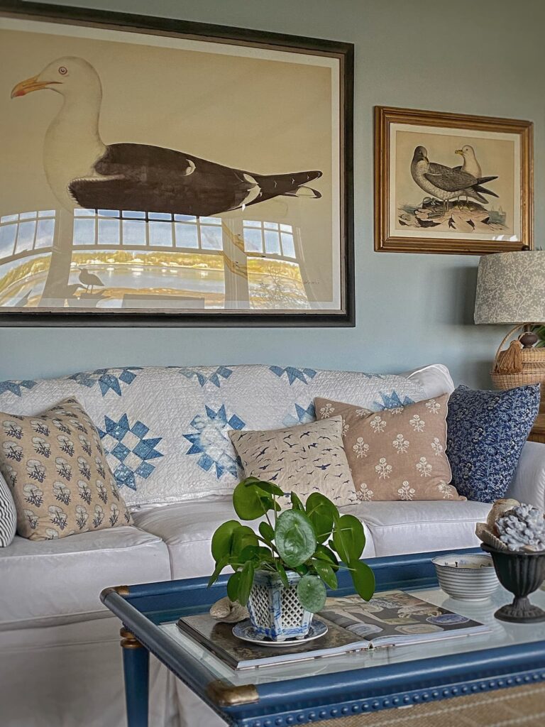 White slipcovered sofa styled for summer with a vintage blue-and-white quilt, patterned throw pillows, coastal bird artwork, and a blue coffee table in a Maine living room.