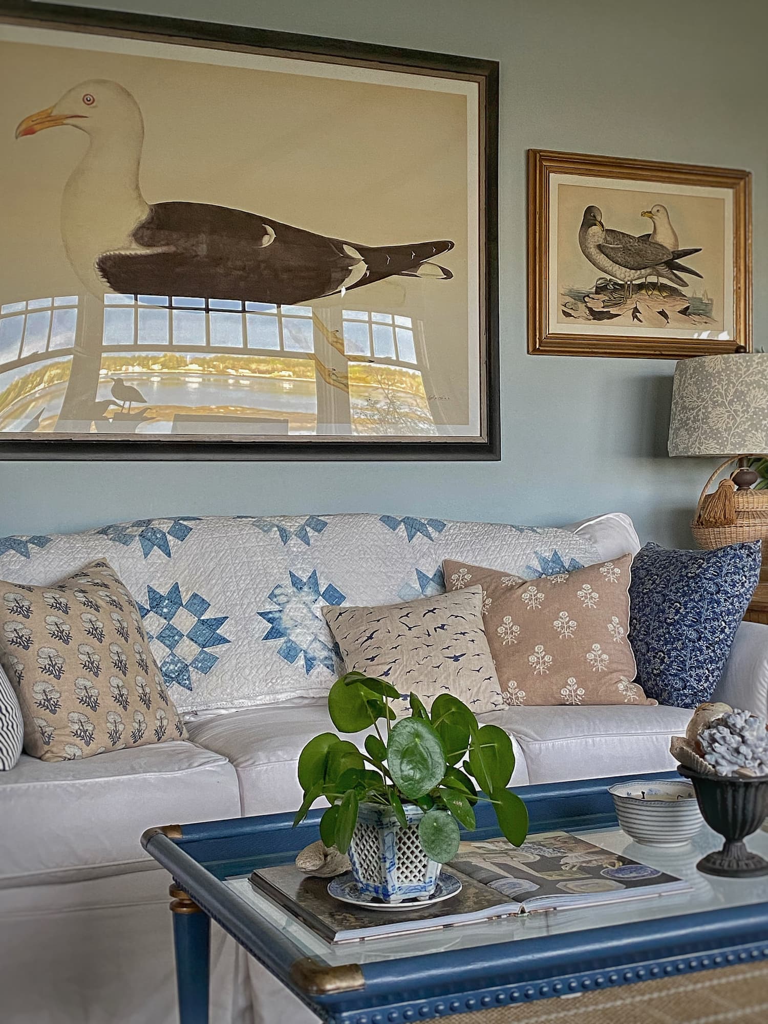 White slipcovered sofa styled for summer with a vintage blue-and-white quilt, patterned throw pillows, coastal bird artwork, and a blue coffee table in a Maine living room.