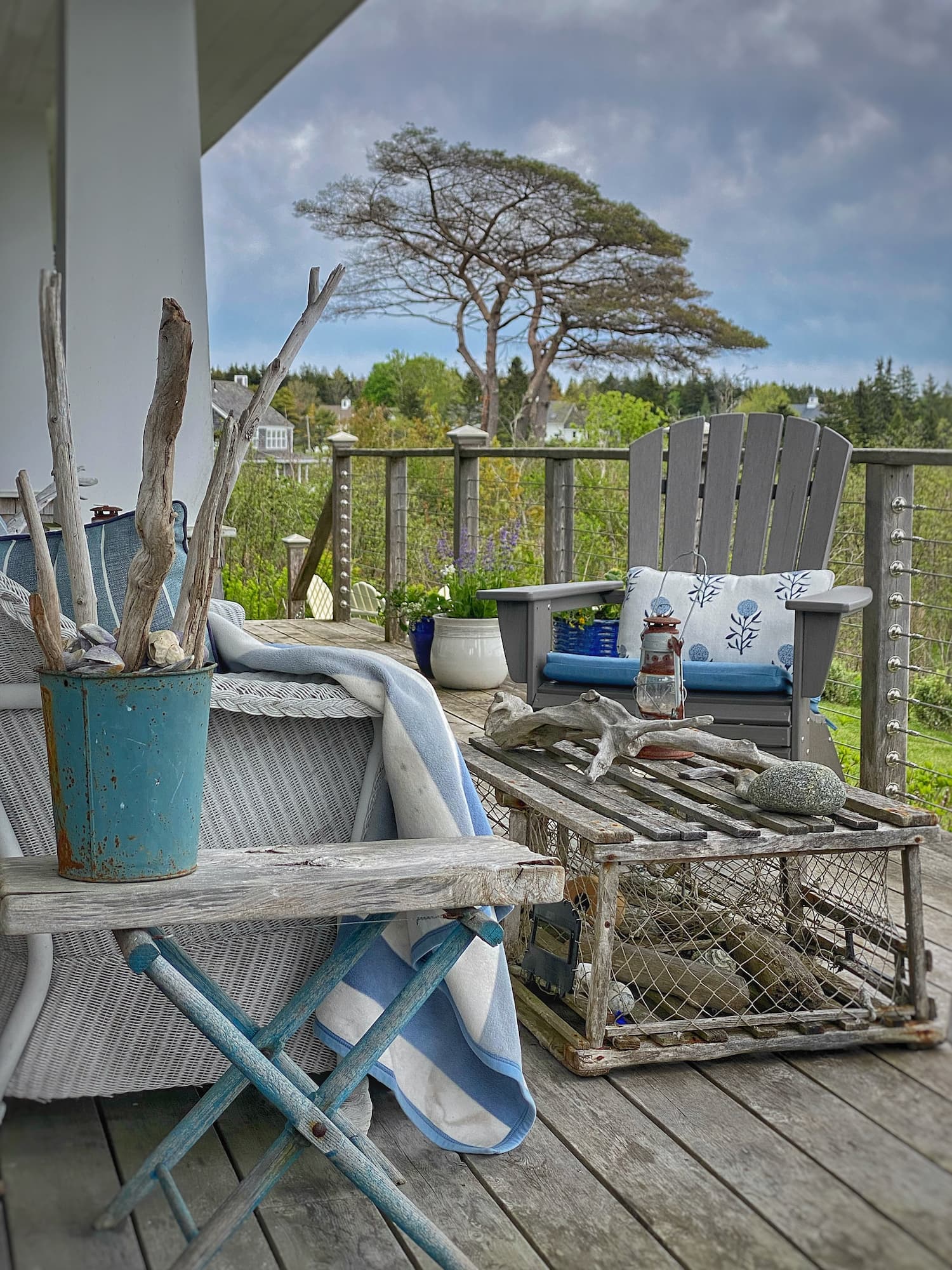 Weathered coastal porch on the Maine coast with an antique lobster trap coffee table, driftwood accents, gray Adirondack chair, wicker settee, and blue-and-white textiles under dramatic cloudy skies.