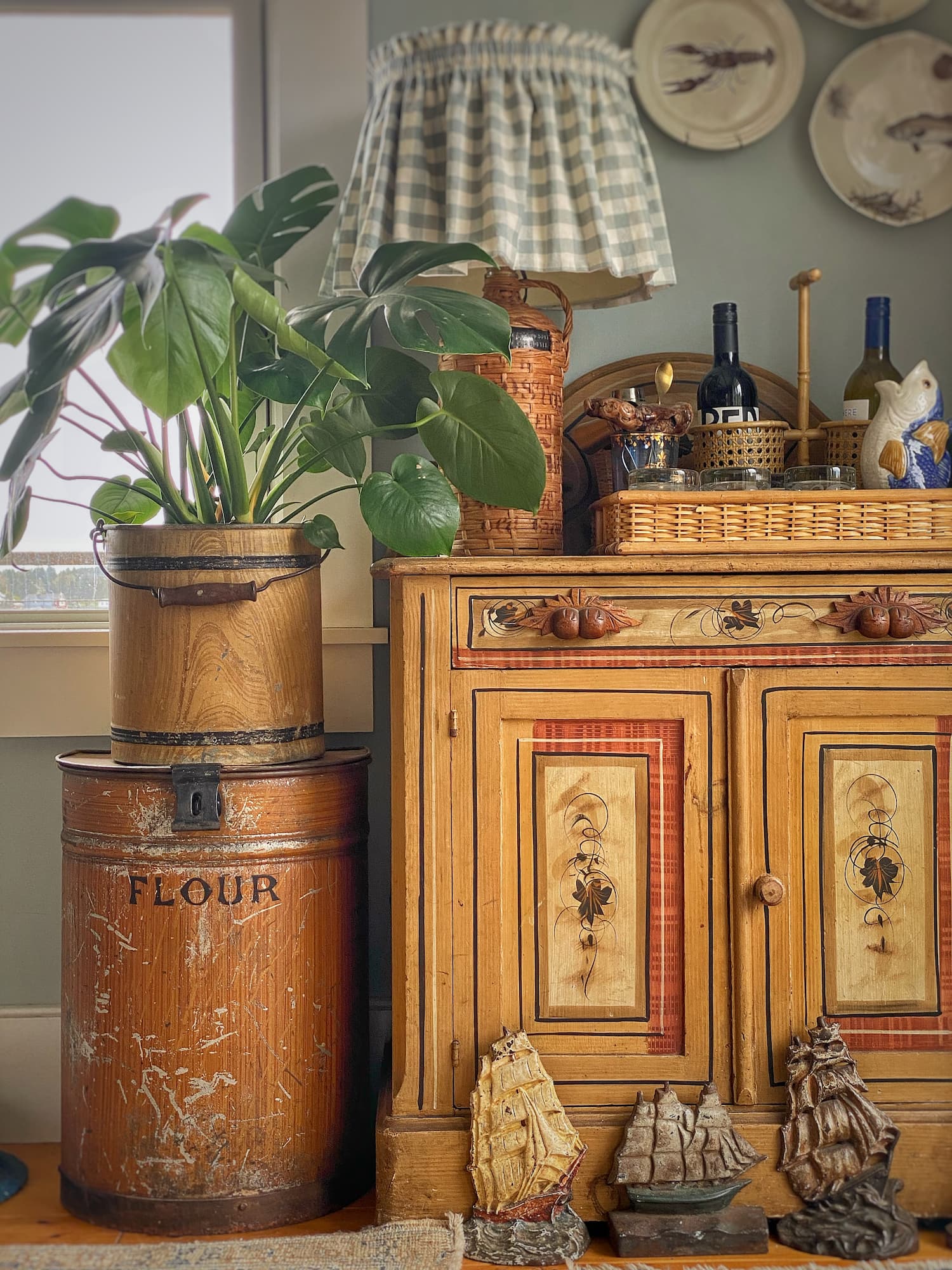 Faux-grain flour bin and painted cottage cabinet styled with plants, faux wicker pieces, and collected coastal decor in a Maine cottage.