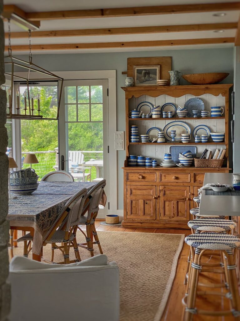 Summer dining room in a Maine coastal home featuring a reclaimed pine farm table, blue-and-white woven bistro chairs, a sisal rug, and a pine hutch filled with blue-and-white Cornishware. Soft blue walls, exposed ceiling beams, and a glass door opening to the porch create a bright, relaxed New England cottage feel.