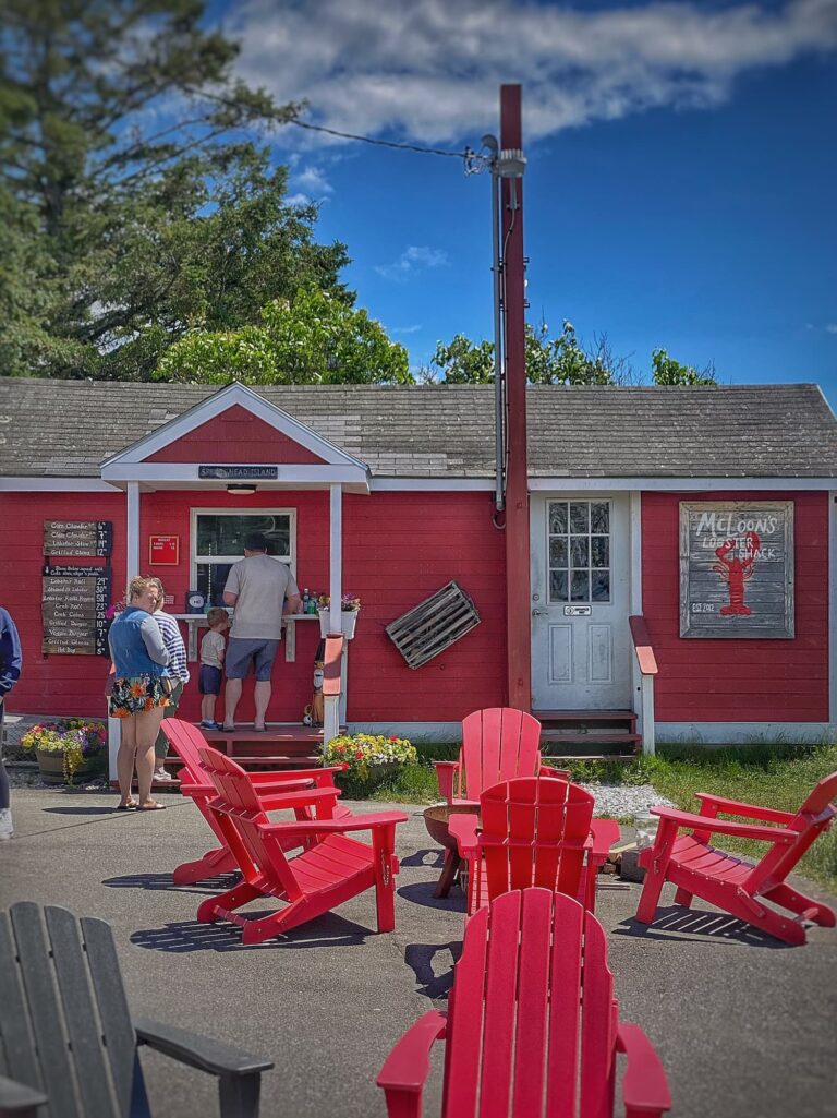 Red Adirondack chairs sit outside McLoon's Lobster Shack on Spruce Head Island, Maine, with the bright red building and blue summer sky creating a classic coastal scene.