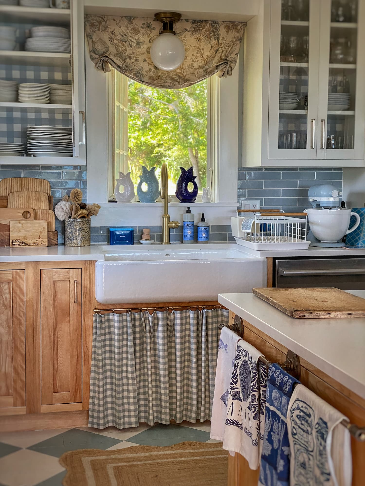 Sunlight streams through a kitchen window above a farmhouse sink, surrounded by white cabinetry lined with blue gingham wallpaper, blue and white dish towels, a café curtain beneath the sink, and everyday kitchen details that create a warm New England cottage feel.