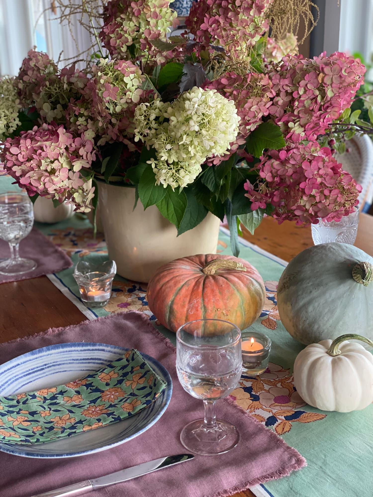 Early fall table set with pink hydrangeas, blue and white dishes, floral linens, candles, and colorful pumpkins.