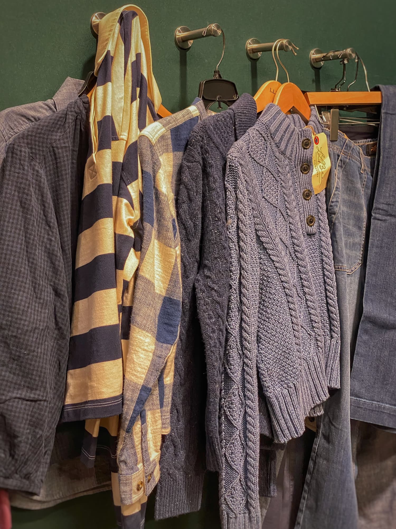 Fall clothing hanging in an L.L.Bean dressing room, including blue cable-knit sweaters, plaid flannel shirts, a rugby shirt, and jeans.