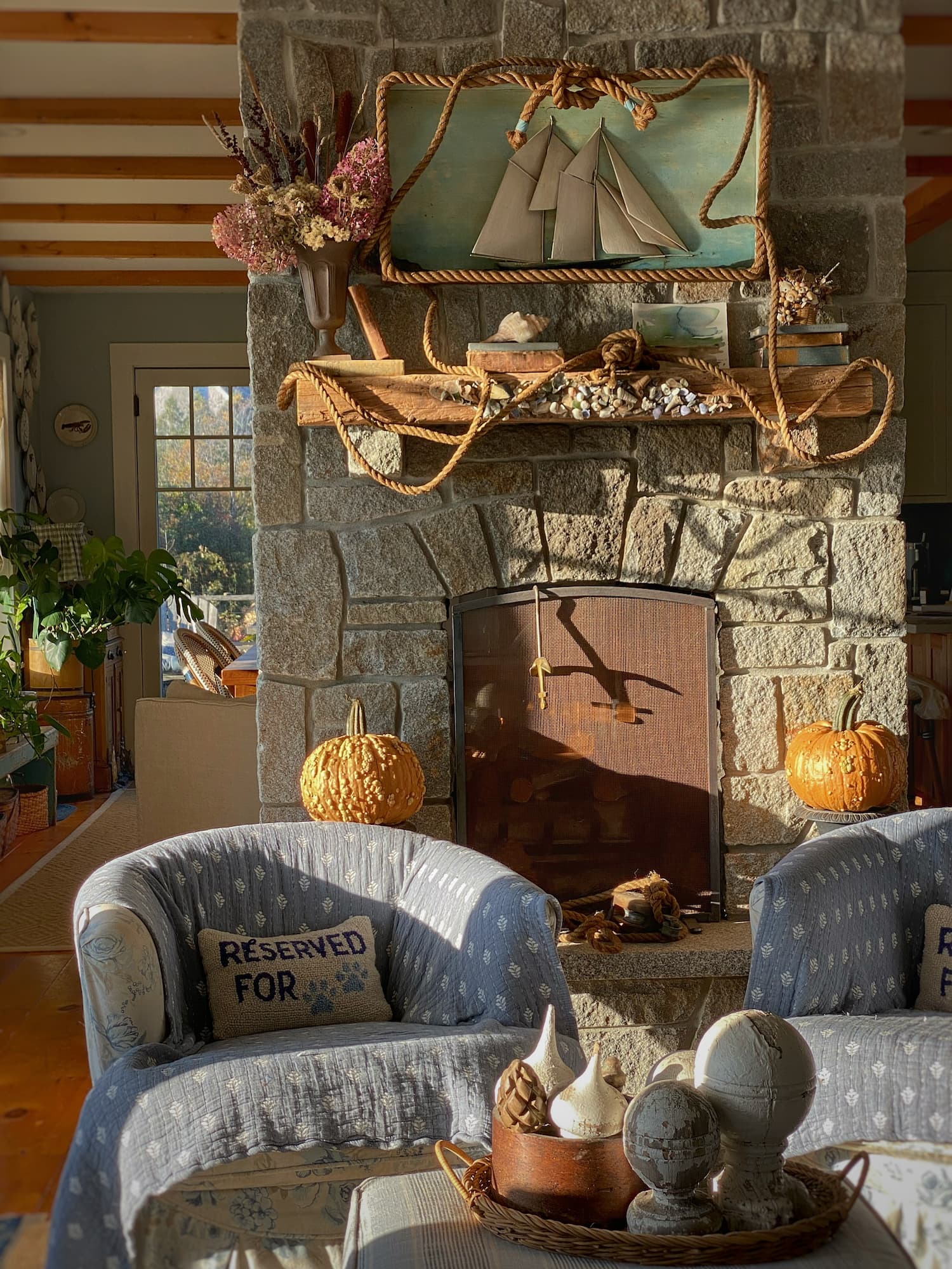 Cozy coastal Maine living room decorated for fall, with golden sunlight on a stone fireplace, caramel-colored pumpkins, dried flowers, nautical mantel decor, and blue slipcovered chairs.